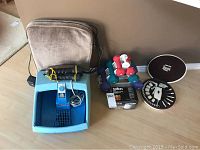 Blue foot massager with a wired control, assorted dumbbells, small black battery charger, black and white flashlights, and sonic jewelry cleaner kit all arranged on floor against wall.