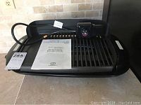 Black electric countertop gourmet health grill with power cord and plug placed on countertop. Instruction/recipe book on top.