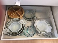 Top view of drawer with white ceramic mixing bowls, clear Pyrex measuring pitcher, round and rectangular glass baking dishes, woven basket, and two wire strainers.