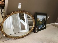 Photo showing the oval gold-colored mirror with ornate frame, acrylic painting, and small framed feather leaning against a wall.