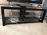 TV stand with black metal rectangular frame, glass top and bottom shelves, placed on carpet with TV and cords on it.