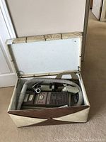 Electrolux vacuum cleaner stored inside open cream vinyl covered chest showing vacuum, hose and upholstery tool inside.