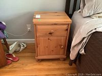 Front view of one pine end table next to bed, showing one drawer and one cabinet door with wooden knobs and rounded bun feet.