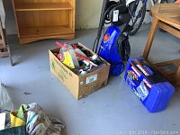 Simoniz S1900 pressure washer unit, blue, next to a cardboard box with assorted cleaning supplies and a hard blue Simoniz accessory case.