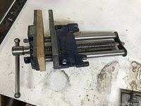 Close view of a 7 inch metal carpenter's woodworking vise with blue finish, wooden jaw inserts, screw mechanism with handle and dual guide rods on a workbench surface with stains.