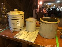 View of 3 pottery crock pots and marble cutting board on wooden table in indoor setting.