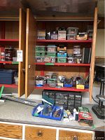 Showcases wooden cabinet with multiple shelves holding boxes and containers labeled with deck screws, lag bolts, roofing nails, hooks, and other fasteners.