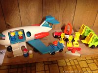 White and blue plastic Fisher Price airplane with wooden character toys and various vehicle and construction toy accessories on wooden surface.