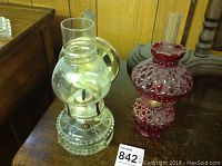 Clear and red glass oil lamps displayed side by side on a wooden surface against a wood-paneled background.