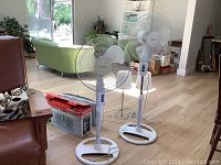 Two white Airworks adjustable height floor fans placed side-by-side on hard wood floor in a bright room, showing overall condition and setup.