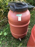 Front view of orange plastic rain barrel with mesh screen cover on top, spigot at the base, standing on grass next to a fence.