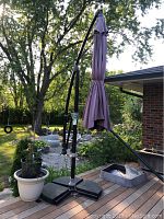 Photo shows the 11-foot offset patio umbrella closed with purple fabric and three black base weights on a wooden deck with surrounding plants.