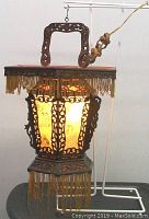 Front and side view of the antique Chinese lantern illuminated, showing carved wooden frame, silk painted panels, and hanging fringes including damage and missing pieces.