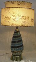 Front view of the table lamp lit, showing the textured two-tiered fabric shade and teardrop glass base with blue and gold horizontal stripes on brass base.