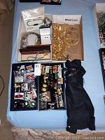 Wide view of the jewelry lot on a bed showing multiple open boxes of necklaces, earrings, and a wrapped black umbrella or pouch.