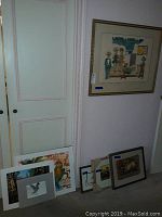 Wide view of several framed and unframed artworks stacked and leaning against wall and door, showing the variety and condition of art pieces.