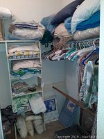 Closet corner showing shelves with folded bedding, pillows, bags, clothes hangers with various hanging garments and boxed storage items.