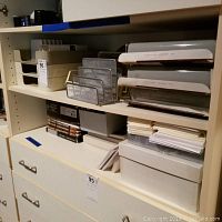 Shelf with metal filing trays, plastic multi-tier letter trays, card storage boxes, and loose papers.
