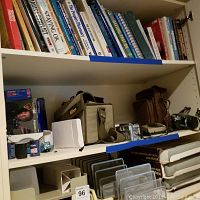 Shelf with manual film cameras, beige and brown camera bags, stacked books above, metal wire office trays below
