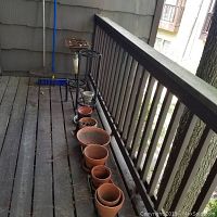 Photo showing nine terracotta flower pots lined up on porch decking, showing various sizes and shapes, some nested inside each other, beside a black metal flower pot stand and a gas grill.