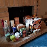 Wide shot showing a variety of candles, a large box of Pine Mountain logs, timers, and matches arranged on a brick fireplace hearth.