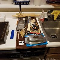Photo of a kitchen counter with a large wooden cutting board loaded with various wooden spoons, rolling pin, knives, and metal kitchen utensils and accessories.