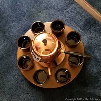 Top-down view of rotary wooden tray holding solid copper fondue pot and eight black ceramic cups arranged evenly around.