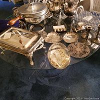 Lot of silver-plated items arranged on glass table including fondue pot, serving tray, candle holders and dishes