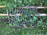 Full view of a black metal gate with ornate fish skeleton design, placed outdoors against a wooden fence with greenery in the background.