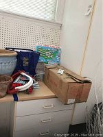 Photo of a table with cardboard box, CD cases, sidewalk chalk, and USA license plate game board leaning against wall.