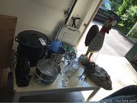 Cookware including large roasting pot, colander, coffee maker, decorative wire chickens, large carved wooden rooster, and resin onion sack decor on table.