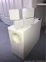 Four white Bose Acoustimass 5 Series II satellite speakers arranged on and beside the white subwoofer bass module. Satellite speakers small cube shape with fabric grille and Bose logo.