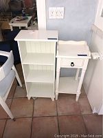 Two white wooden furniture items side by side: a three-shelf accent shelf and a side table with drawer and lower shelf, showing overall condition and style.