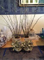 Two tall bamboo-style silk plants in square beige pots displayed side by side on a floor with two smaller leafy silk plants with flowers in front of them.