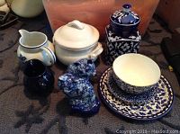 Blue and white antique English potpourri vase, two cats figurines, cream and blue canister, rustic white canister, blue and white plates, bowl, and small pitchers arranged on a surface