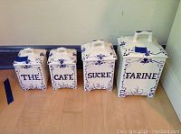 Four Jay Willfred blue and white ceramic kitchen canisters arranged by size with French labels THE, CAFE, SUCRE, and FARINE.
