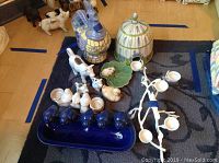 Photo of clustered ceramic items including the Food Network cow tea set with cups and tray, white bird candle holders, ceramic bunny, and duck figurines arranged on a floor rug.