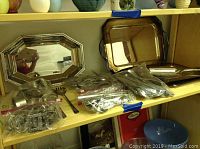 Silver plated serving trays with reflective surfaces shown on shelf, some flatware and other items bagged in plastic also visible