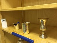 Three tableware items on a wooden shelf including a small cobalt enamel dish, a silver plated beaker, and a gold washed sterling silver goblet.