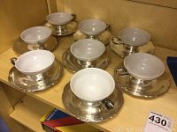 Wide view of seven porcelain cups with silver plated overlay paired with seven matching saucers on a wooden shelf. Two cups have minor repairs.