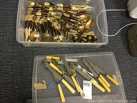 Top view showing clear plastic storage bin filled with brass and wood flatware including knives, forks, and spoons, plus vintage knives with yellow Bakelite handles in a separate black container.