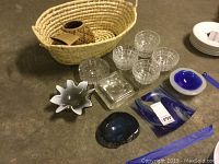 Photo of large woven basket with small Zulu basket inside, crystal berry bowls arranged on floor, and two art glass ashtrays, plus some white plates on the side.
