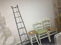 Full view of the metal pie rack, wooden stool, and two country-style dining chairs against white wall.