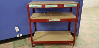 Front view of red metal shelving unit with three shelves, showing stains on the shelves and labels on the top shelf edge.