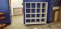 Front view of white cubby hole unit with 15 compartments on wheels in a room with tiled floor and blue walls.