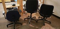 Three computer office chairs in a room with wooden shelving, showing their backs and bases. Chairs are dark colors with adjustable armrests and wheels.