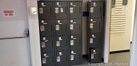 Bank of twelve tall gray metal lockers with three vertical columns and four horizontal compartments each with lock handles, shows multiple stickers on locker doors.