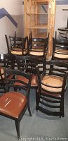 Set of twelve stacked black wooden chairs with brown cushioned seats in a storage room, some cushions facing upwards showing wear and tears.
