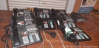 Photo showing eight Norstar telephone handsets arranged on a table, seven M7310 and one M7208 model, black multi-line office phones.