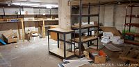 Two metal-framed shelving units in basement setting with surrounding miscellaneous items. One short and one tall unit with particle board shelves.